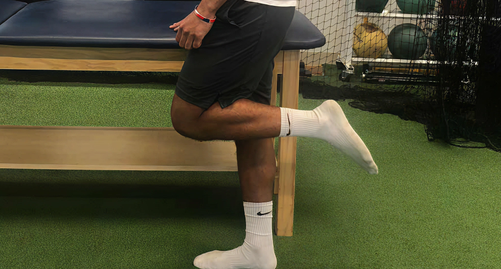 A side view of a person standing on one leg with the other bent back at the knee, performing a hamstring curl or stretch in a gym with green turf flooring.