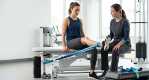 A woman sitting on a treatment table performs a leg extension with a resistance band, guided by a physical therapist in a bright, modern clinical gym setting.