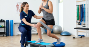 A physical therapist in blue scrubs assists a patient performing a banded lunge using a step platform and balance disc in a bright, well-equipped clinic.