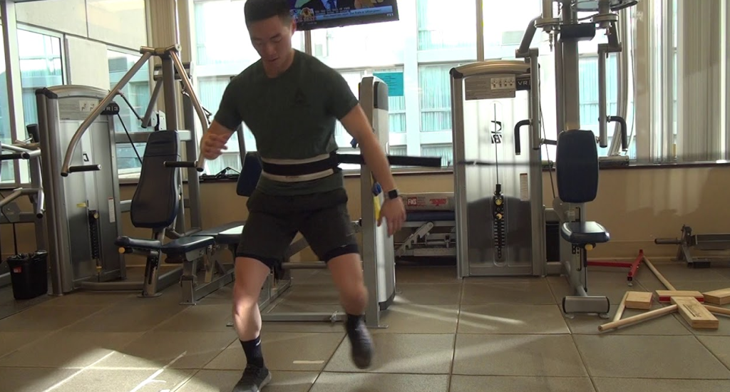 A person wearing a workout belt and gym attire performs lateral jumps in a fitness center. Resistance machines and gym equipment are visible in the background.