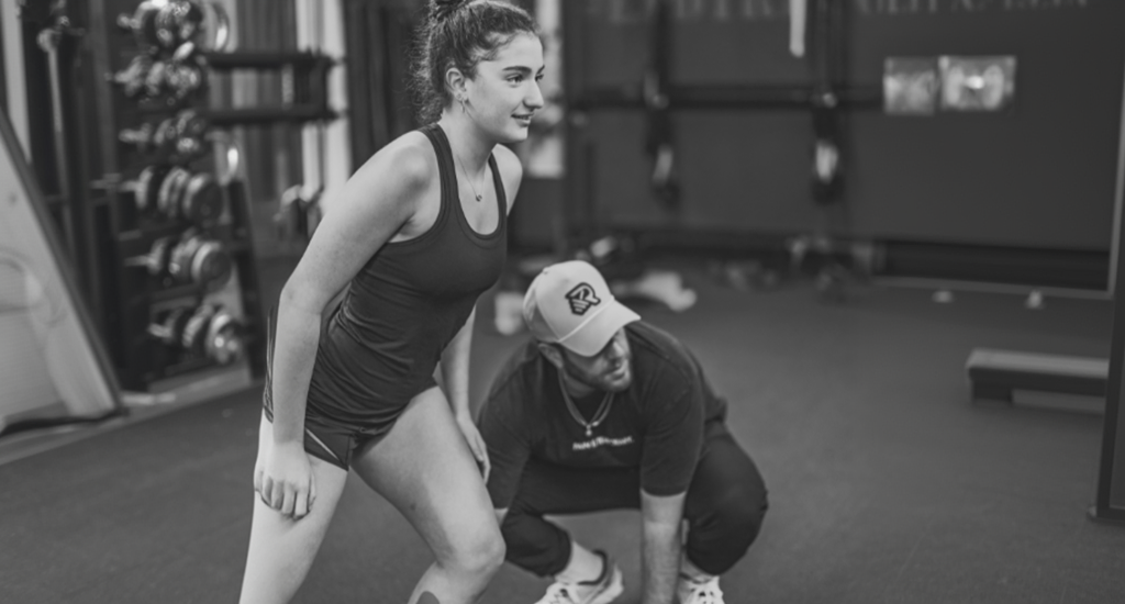A black-and-white photo of a young woman in athletic wear focused during a workout, while a trainer in a baseball cap crouches beside her to guide her form.