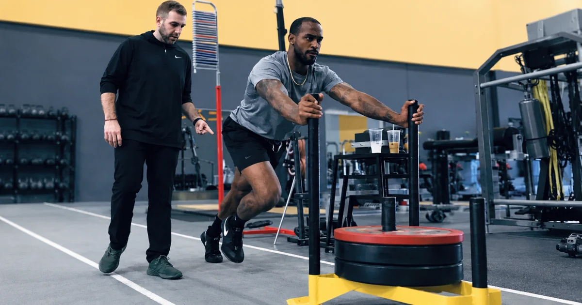 An athlete doing a weighted sled push exercise on a track while a physical therapist points and guides him.