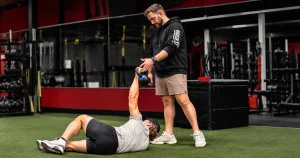 A fitness trainer guiding an athlete through the initial floor phase of a Turkish Get-Up exercise using a kettlebell on an indoor turf.